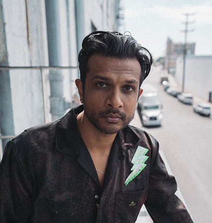 A photo of Utkarsh Ambudkar standing on a balcony, with city street and parked cars behind him.