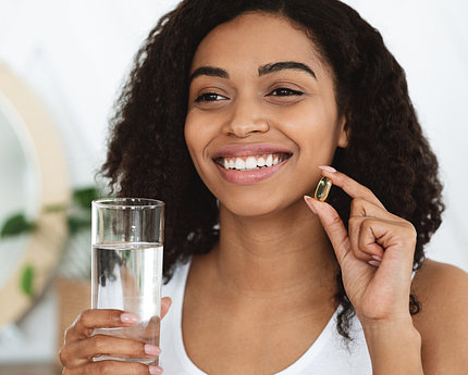 A smiling young African American woman holds up a yellow capsule in one hand and a glass of water in the other.