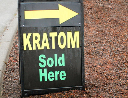 On a gravel path in Oregon, a sign with large, bright letters reads: Kratom Sold Here, with a large arrow