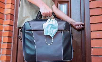 A man's hand is wrapped around his home's door handle while his other hand holds a black briefcase and surgical mask.