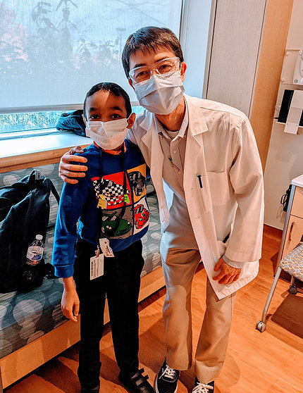 Dr. Hsieh (pronounced "Shay) stands with his arm around Caesar, who are both masked, in Caesar's room at the Clinical Center, autumn 2021