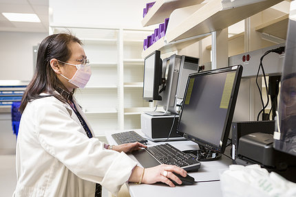 Woman in lab coat stands at computer screen while using mouse attachment.