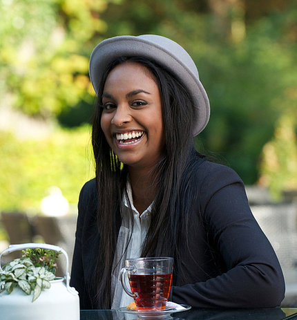 An African-American woman wearing brimmed hat sits at table, smiling widely, with cup of black tea on table, trees behind.