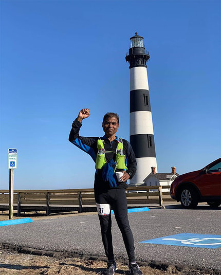 Bhattacharyya stands triumphant in running gear, one fist in the air, in front of lighthouse and bright blue sky.