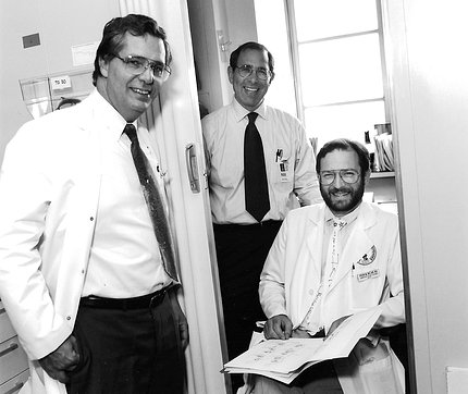 Three men, two in lab coats, smile at camera from doorway to laboratory