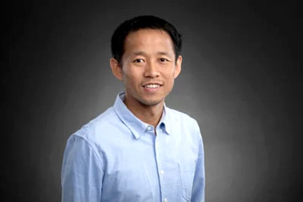 Zhou poses against a gray background. He is smiling and wearing a blue shirt.
