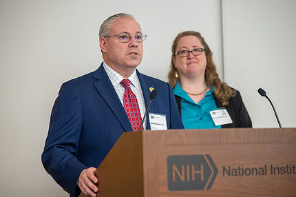 Grossman speaks at a wooden podium with an NIH logo, with Shivas standing behind and to the right.