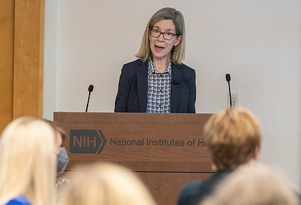 Criswell speaks at a wooden podium with an NIH logo on its front. She wears glasses and a black suit jacket, with a black and white patterned shirt.