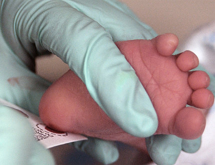 Gloved hand holds a newborn infant's foot while performing a heel prick rest.
