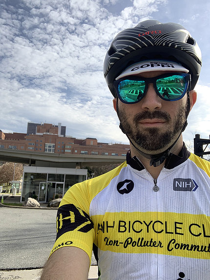 Velez, in NIH Bike Club shirt, helmet and sunglasses, stands near Clinical Center
