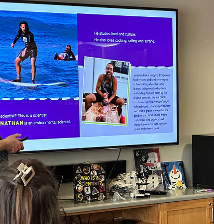 A video screen shows pages from Gehl's book: Nathan, an environmental scientist, is shown surfing, which is listed among his hobbies.