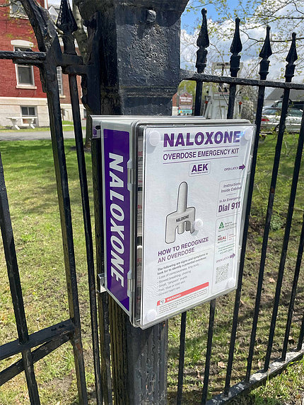 A portable box containing naloxone doses hangs on black fence on street