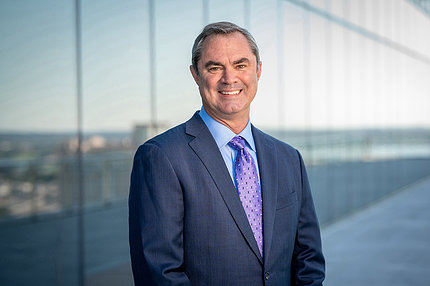 Smith stands outdoors in front of a mirrored wall reflecting a city skyline. He is wearing a suit and purple tie and is smiling.