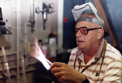 Dehn holds a thin glass cylinder to a stream of white-hot flame. He wears a striped shirt and safety glasses, with another headpiece propped on his forehead.