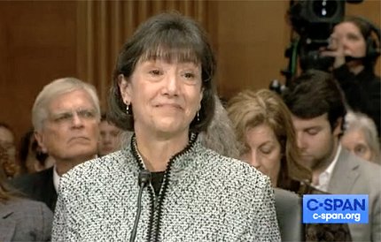 Screenshot of Dr. Bertagnolli at a Senate hearing during her confirmation process. She stands at a podium with a microphone, with a crowd of viewers in the background that includes a camera and videographer. There is a blue C-SPAN logo in the bottom right corner of the image.