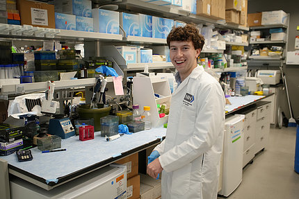 King, wearing a white lab coat and standing in front of a lab bench with shelves of scientific instruments above him.