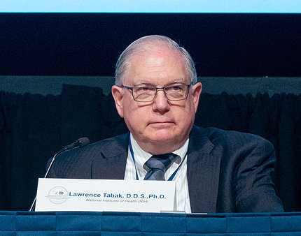 Tabak sits, with name placard, at a conference table.