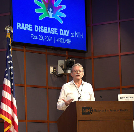 Hickman speaks at a podium. A screen behind him shows the Rare Disease Day 2024 logo.