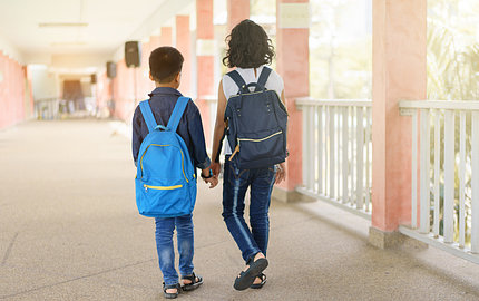 Two children wearing backpacks walk hand-in-hand away from the camera.