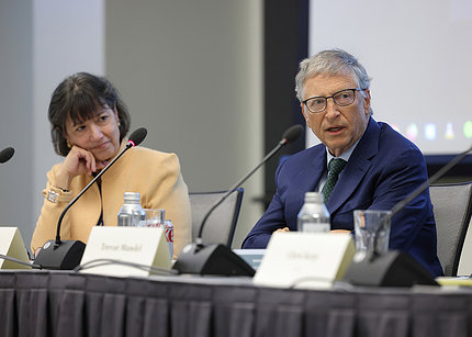 Gates and Bertagnolli seated at a long table. Bertagnolli, to the left and slightly out of focus, listens to Gates, who speaks into a microphone while looking to the right and out of the frame.