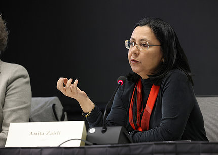 Zaidi, seated, speaks into a microphone and gestures with her right hand.