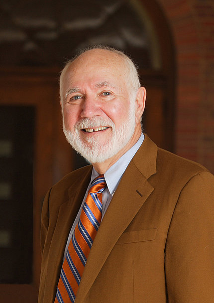 Slavkin smiles at the camera. He is bearded and wears a tan suit jacket and orange, blue and white-striped tie.