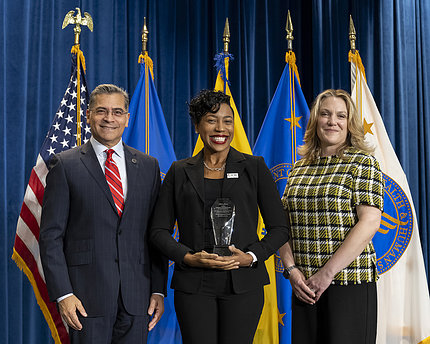Jackson, smiling broadly and holding 8CRE's award, stands between Becerra and Palm. Five flags are lined up behind them, including an American flag on the far left, with a dark blue curtain behind the flags.