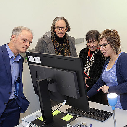 Woman at computer monitor gestures toward screen as man and two other women look on.