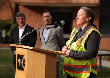 A woman in a fluorescent vest speaks into a microphone at a podium bearing an NIH logo. Two men look on in the background.