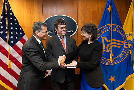 Becerra and Monica Bertagnolli, smiling and shaking hands.Bertagnolli's husband holds a book, and smiles.