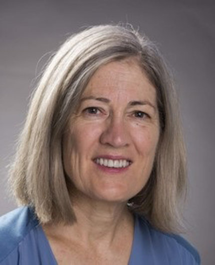 A smiling Moran poses against a gray background. She wears a blue shirt and has shoulder-length gray hair.