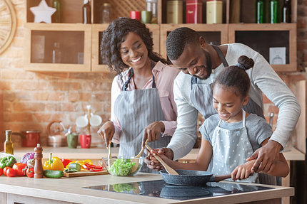 Black father watches young black daughter stir pan on cooktop as black mom looks on, mixing salad.