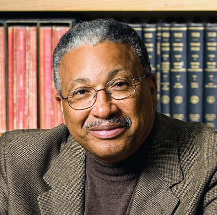 Black man smiling, wearing jacket and glasses, with bookshelves behind him