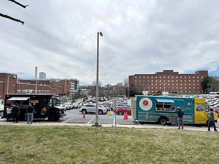 Two large food trucks parked along the sidewalk at the end of a parking lot. 