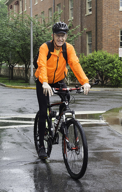 Collins in helmet and on bicycle pedals along wet pavement in front of Bldg. 1.