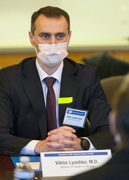 Ukraine's health minister wearing a mask, sits with hands folded at table in CC medical board room, listening intently to NIH staff briefing.