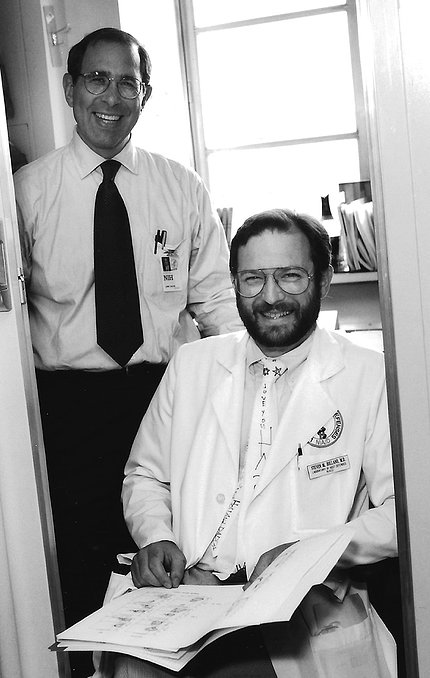 Black & white photo of a smiling, bearded Holland sitting, with Gallin smiling, standing next to him in an office, in 1996