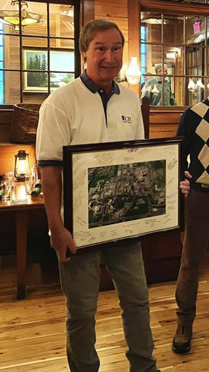 McLean, wearing a white CIT polo shirt, holds a large framed photo showing an aerial view of the NIH campus. The image is surrounded by a white border that has been filled with signatures and well wishes.