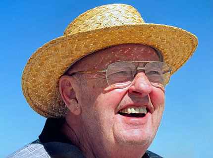 Dehn smiling against a blue sky. He wears a straw hat and glasses.