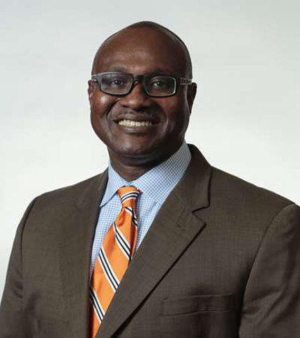 Egede posing against a white background. He is smiling and wearing glasses, a brown suit, and blue shirt and orange-striped tie.