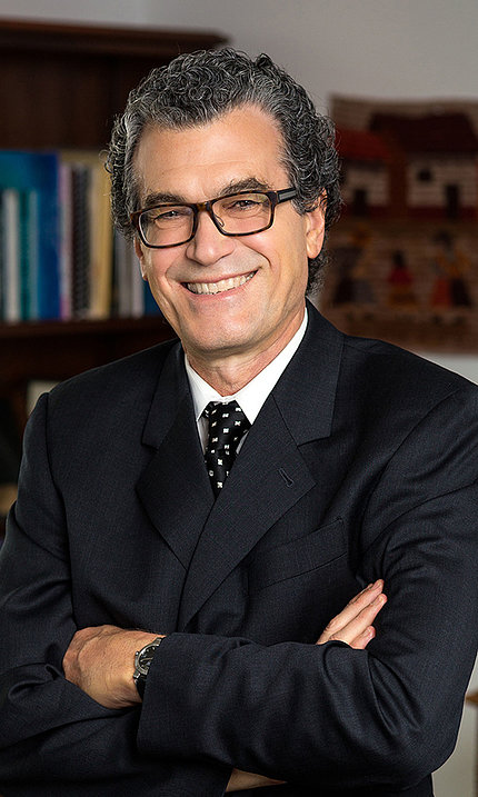 Pérez-Stable smiles with his arms folded. There is a bookshelf in the background. He wears a black suit, white shirt and black tie with a white pattern.