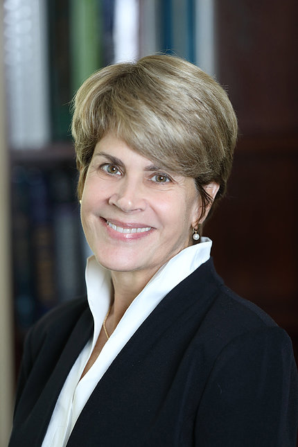 Dr. Tucci smiles against a bookshelf. She is wearing a white shirt and black suit jacket.