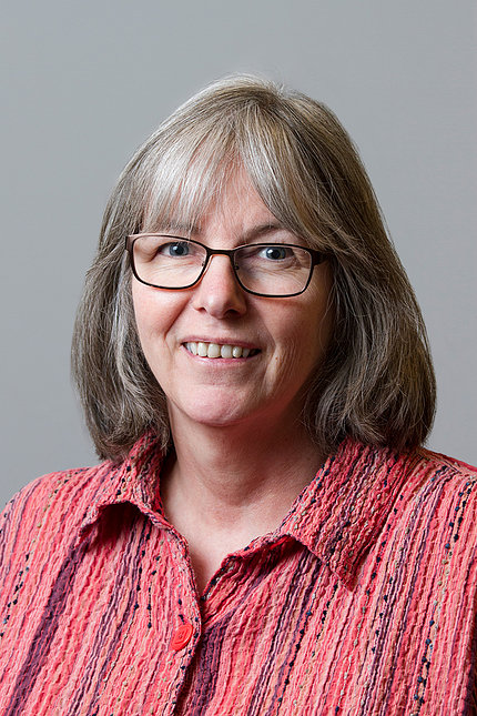 Headshot of Mcguirl, who smiles against a light gray background. She wears glasses and a red patterned shirt.
