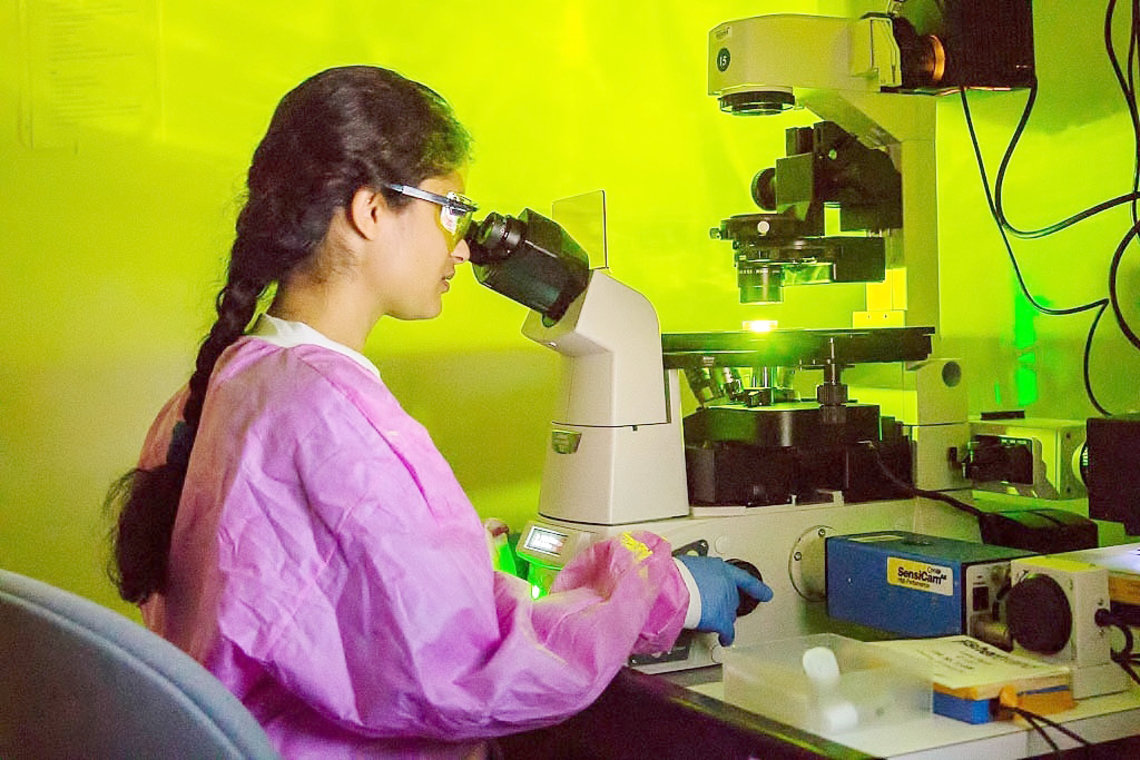Woman in pink lab gear and blue gloves, seated at microscope