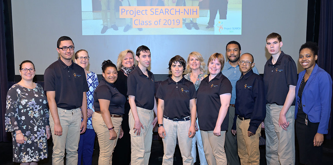 The 7 Project SEARCH grads pose with their instructors on the Lipsett Amphitheater stage.