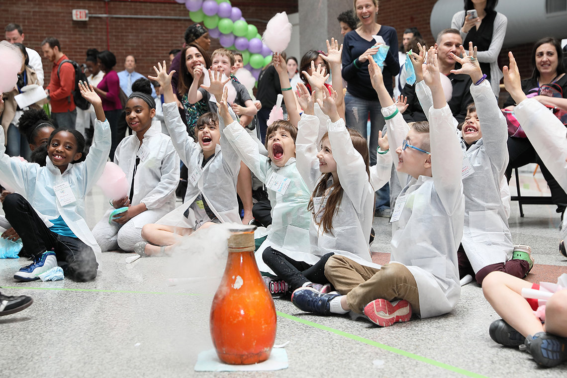 Smiling kids throw hands up and cheer as they watch an orange bottle fizz during a science experiment.