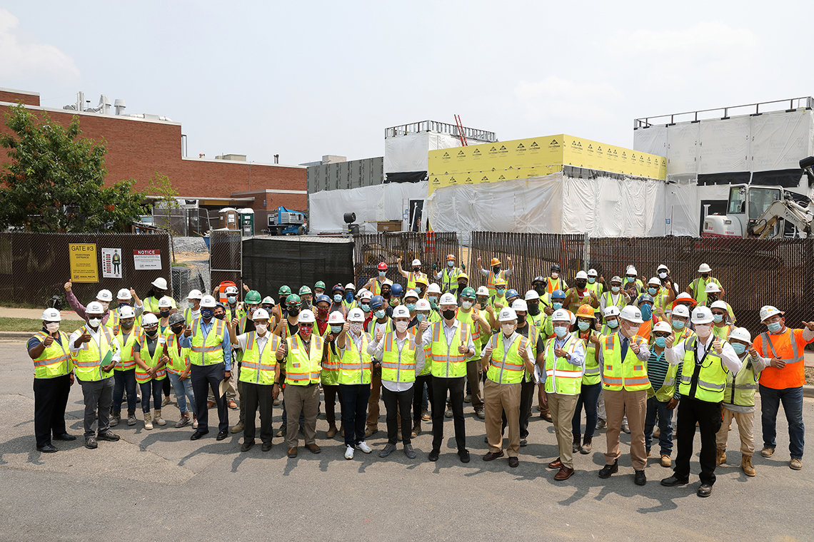 Large group in helmets, masks, and safety vests gives thumbs up in front of construction site.