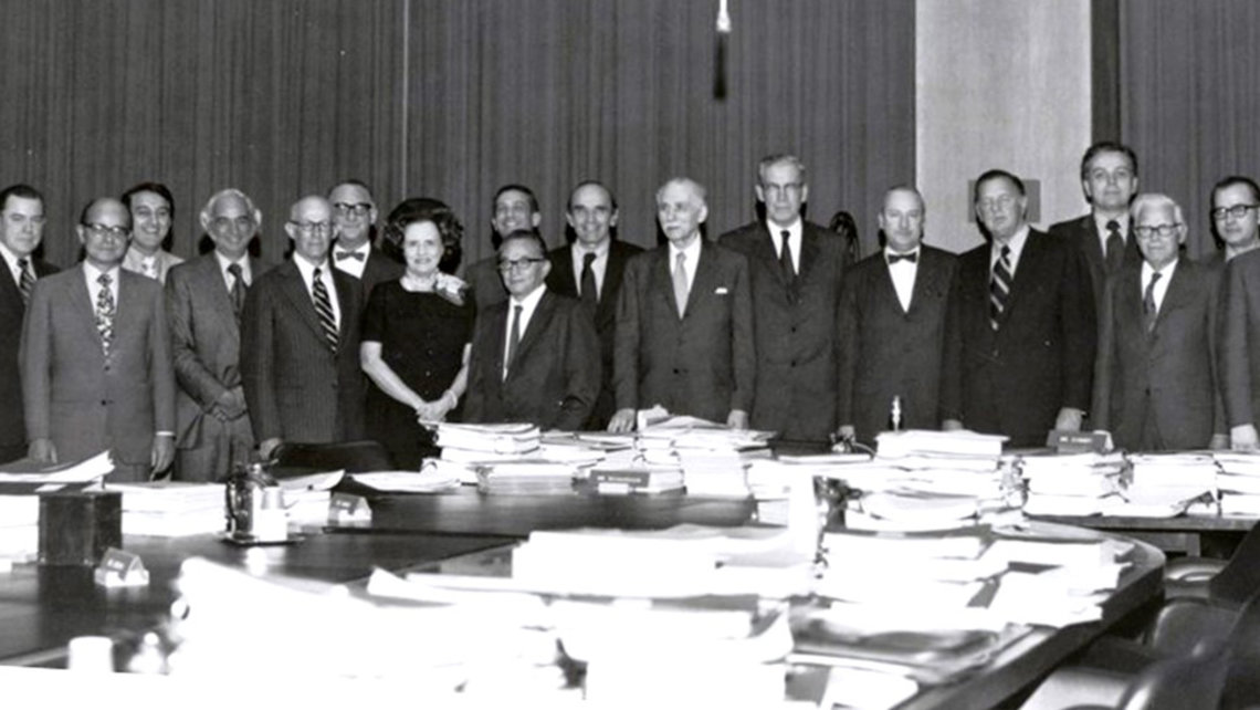 A group of men and Mary Lasker pose for a group photo in front of a table with stacks of paper