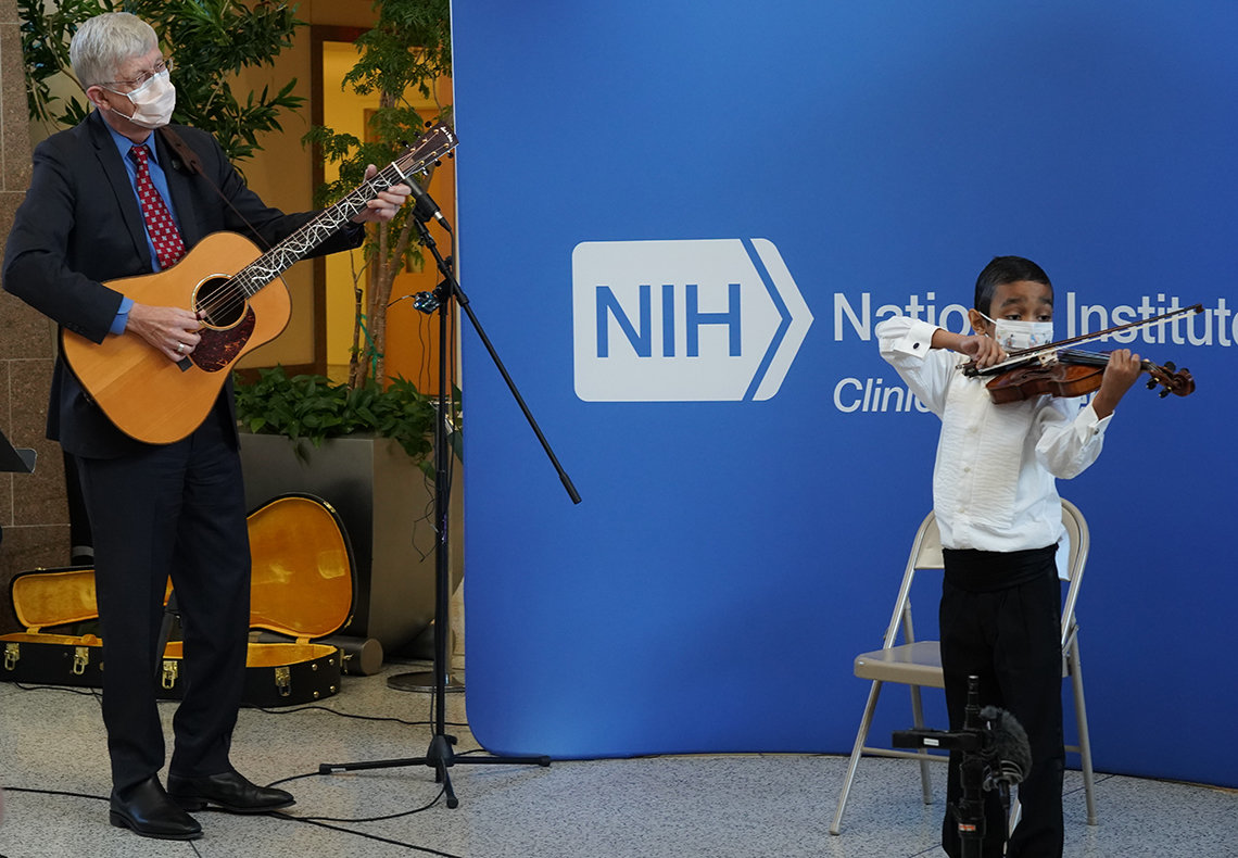 Collins stands playing guitar looking over at Sant playing violin, in front of blue NIH banner in the atrium.