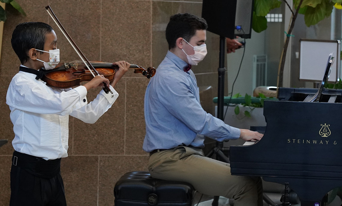 Caesar, in white shirt and mask, holds bow to violin in side view that shows Robert Masi with hands on piano keys in the Clinical Center atrium.